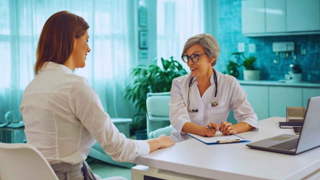 A female patient is consulting with an older female doctor, who is wearing a white coat and stethoscope, sitting across a desk with a laptop and clipboard. This scene illustrates a medical consultation, potentially for an OBGYN checkup for PCOS. 